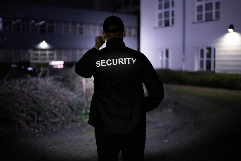 A uniformed security guard on patrol at night, flashlight in hand, back turned toward the camera